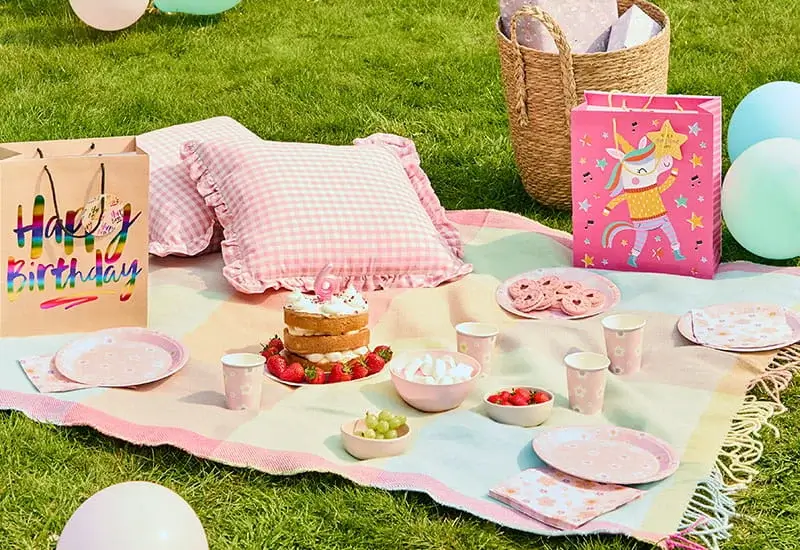 Pastel birthday picnic on a garden lawn with a birthday cake, pink tableware, balloons, gift bags, cushions, and party treats.
